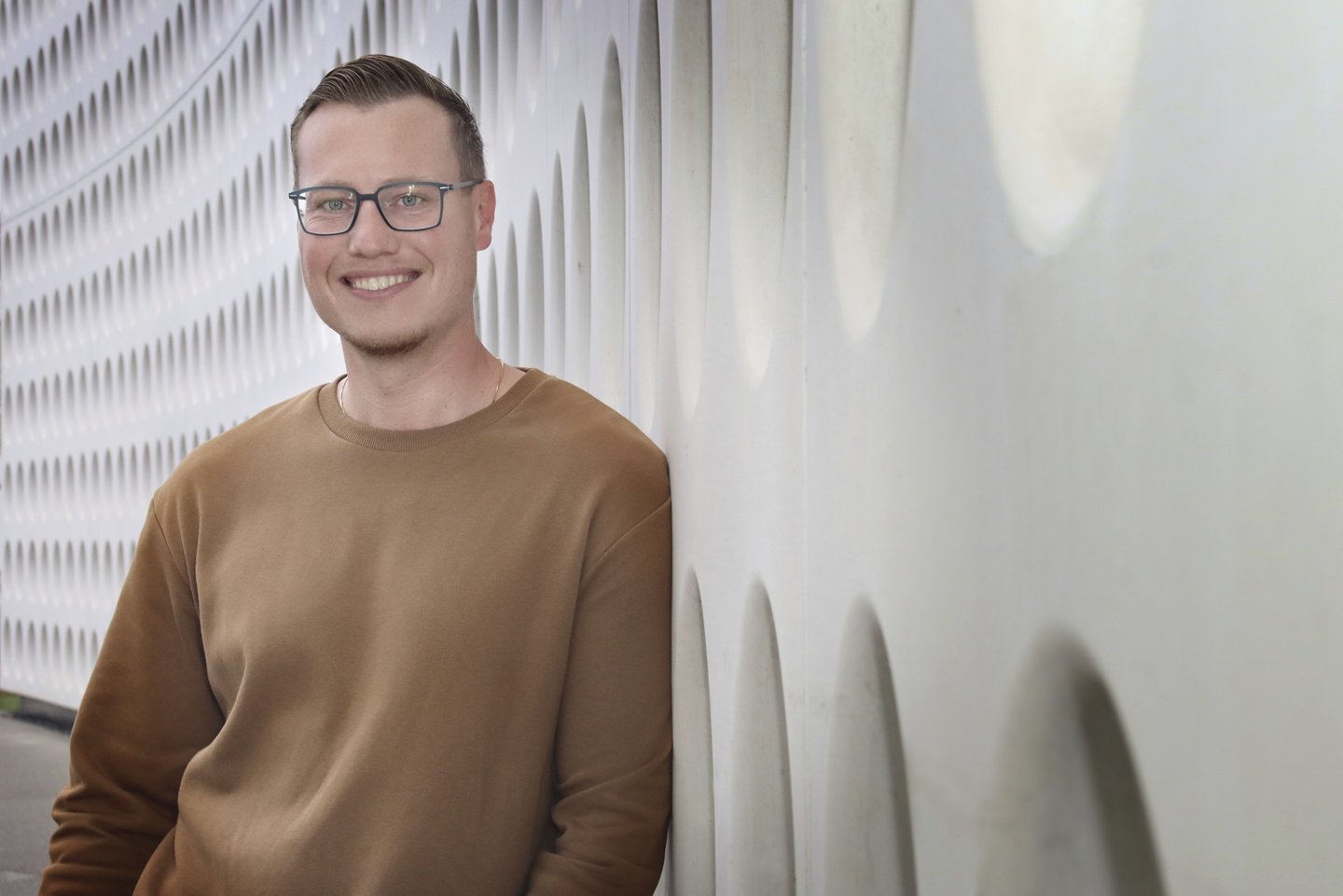 Junger Mann mit Brille, kurzen Haaren und braunem Pullover, lehnt sich an die Außenwand der Silhouette Group in Linz.