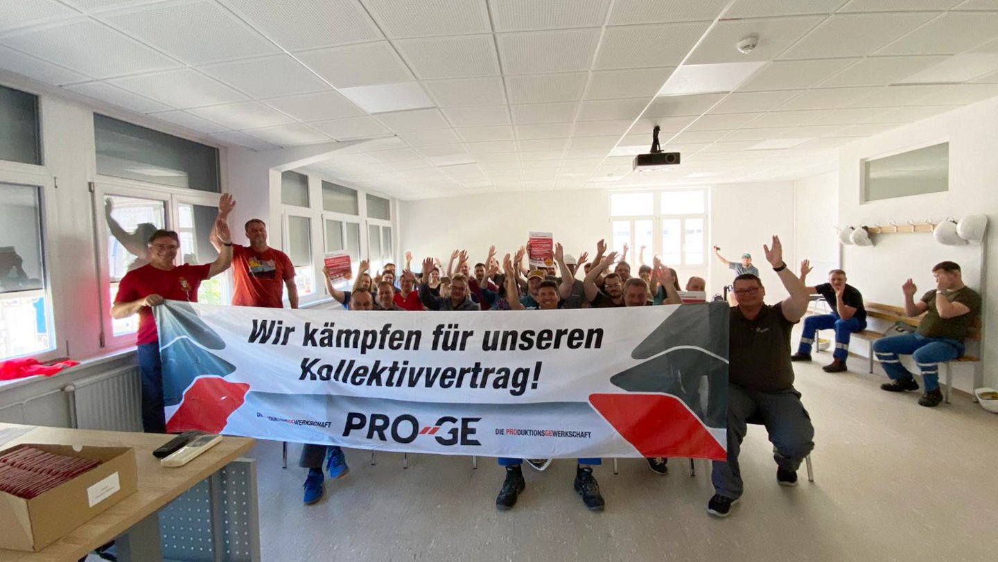 Teilnehmer:innen der Versammlung, sitzend, mit Transparent "Wir kämpfen für unseren Kollektivvertrag!"