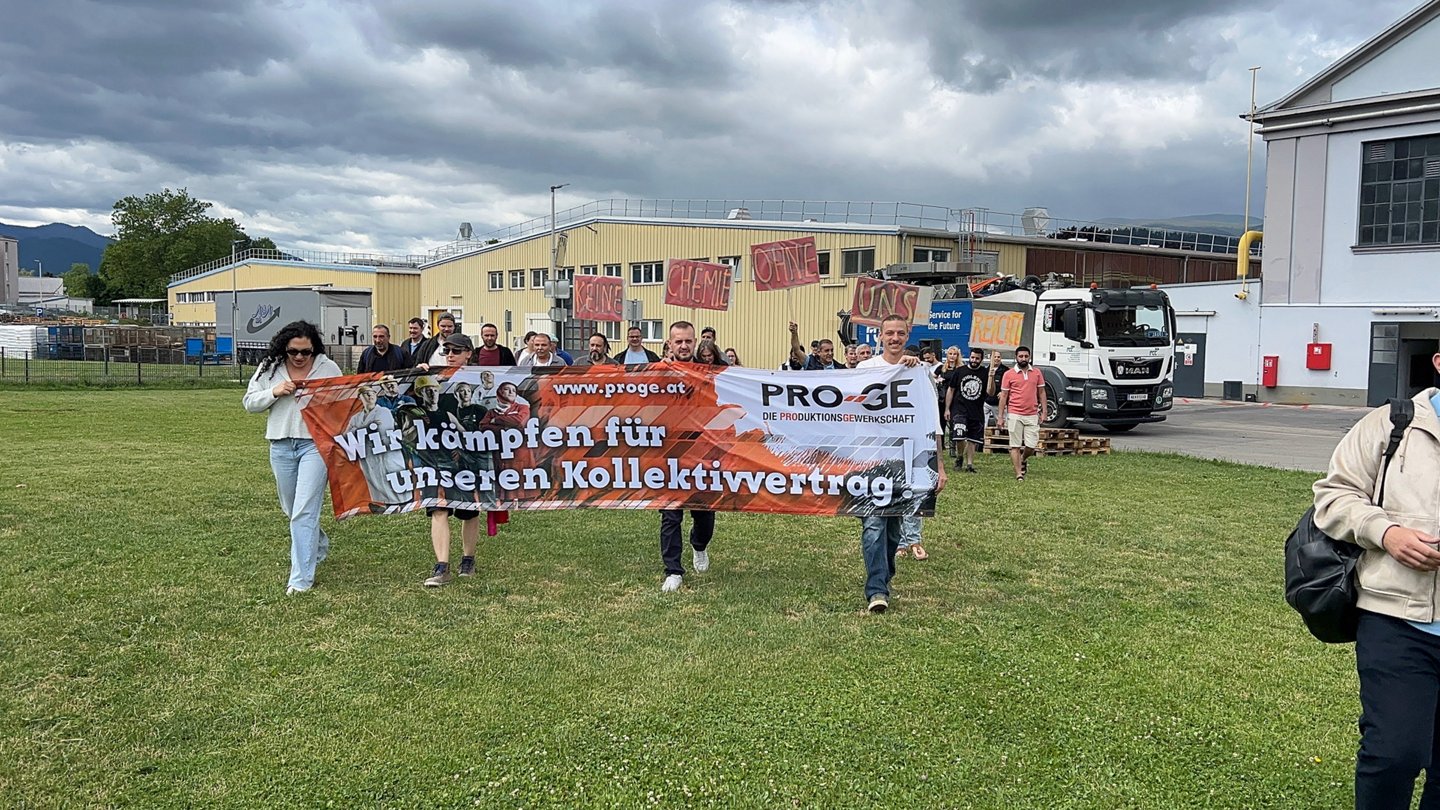 Beschäftigte ziehen mit einem Transparent "Wir kämpfen für unseren Kollektivvertrag!" und Schildern "Keine Chemie ohne uns" über eine Wiese vor dem Werksgelände
