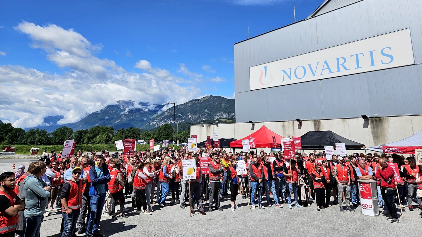 Beschäftigte mit Protestschildern vor einer Werkshalle mit großem Novartis-Logo, im Hintergrund Berge