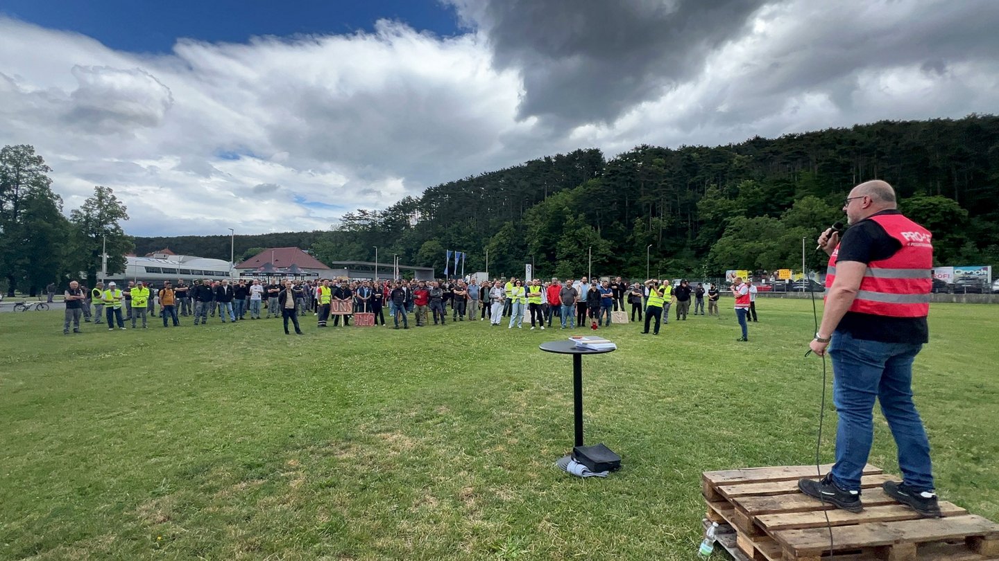 Ein Redner mit roter PRO-GE Warnweste spricht vor den Beschäftigten, die sich auf einer Grünfläche auf bei der Werkseinfahrt versammelt haben