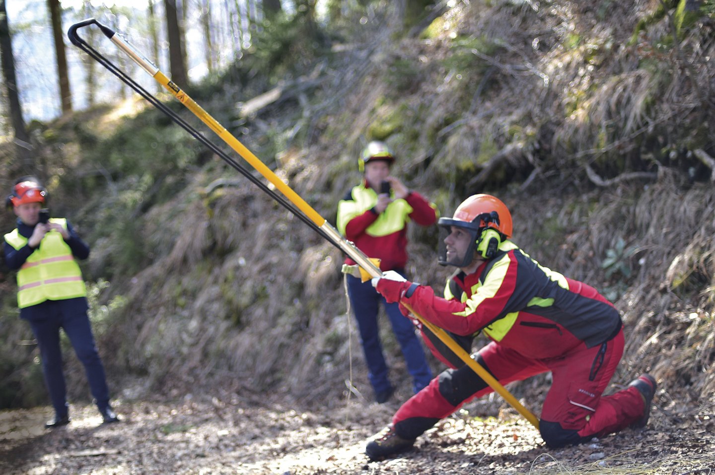 eine Gruppe Arbeiter stehen im Wald, ein Arbeiter mit Helm hält eine Metall-Stange, dahinter stehen zwei Männer, die mit ihren Handys filmen