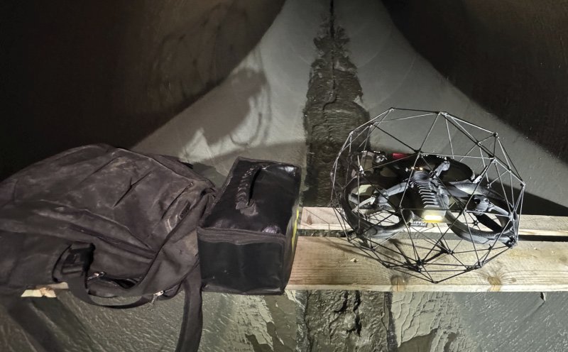 Eine Drohne in einem Schutzkäfig liegt auf einem Holzbrett in einem engen Tunnel. Neben der Drohne befinden sich eine Tasche und ein Rucksack. Die Drohne ist für den Einsatz in gefährlichen oder schwer zugänglichen Bereichen der Bau- und Bergbauindustrie konzipiert.