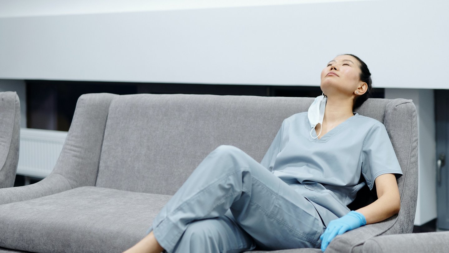 Eine erschöpfte Pflegerin ruht sich auf einem grauen Sof aus