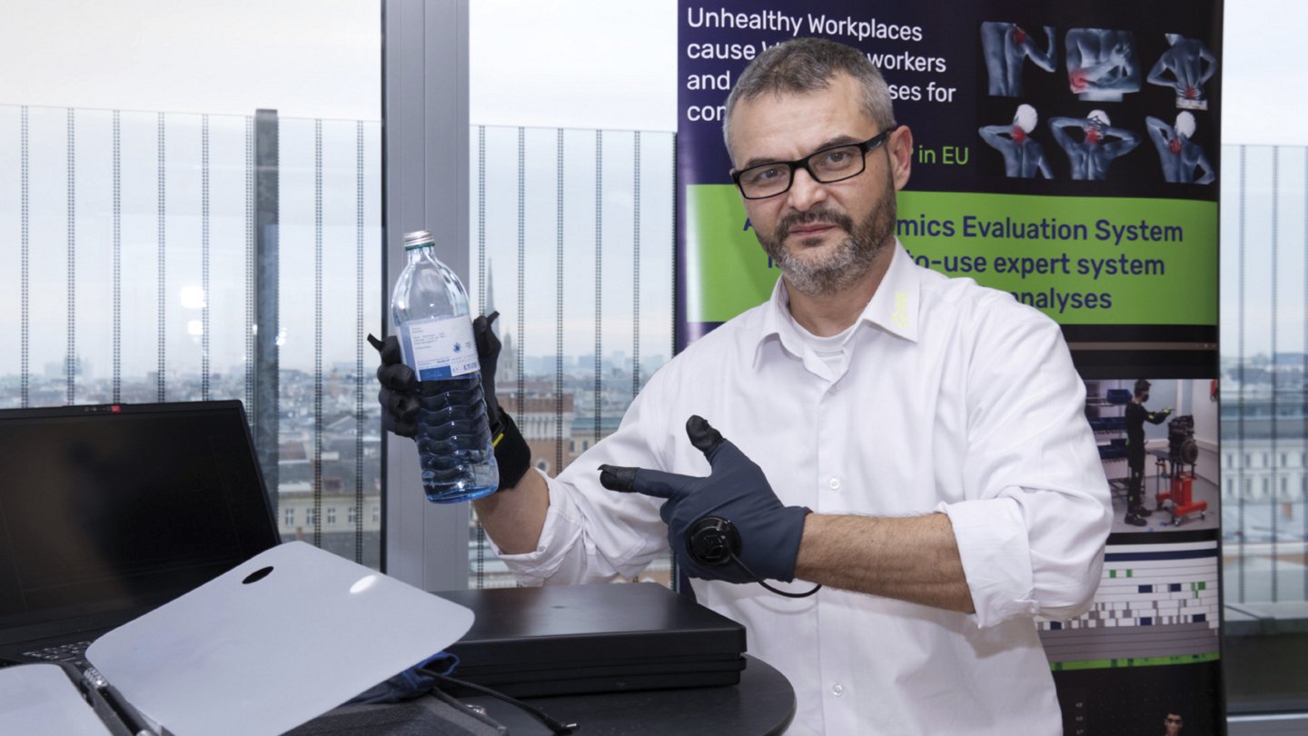 Ein Mann mit Brille und kurzem Bart steht vor einer großen Fensterfront mit Stadtblick und präsentiert eine ergonomische Demonstration. Er trägt schwarze Handschuhe mit Sensoren und hält eine Wasserflasche in der Hand. Auf dem Tisch vor ihm stehen zwei geöffnete Laptops. Im Hintergrund ist ein Banner mit anatomischen Illustrationen und dem Schriftzug „Ergonomics Evaluation System“ zu sehen.