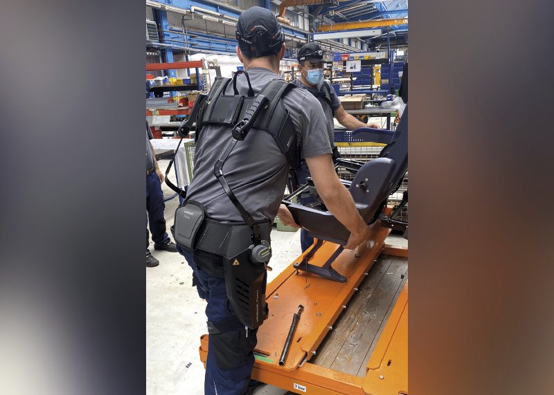 Ein Arbeiter trägt ein Exoskelett zur Unterstützung seines Rückens und ist in einer industriellen Umgebung zu sehen. Der Fokus liegt auf dem Rücken des Arbeiters, der ein schweres Objekt auf einen Transportwagen hebt. Das Exoskelett ist um seinen Rücken und seine Hüfte geschnallt, mit einer Vorrichtung an den Beinen und am Rücken, die Bewegungen unterstützt und Belastungen reduzieren soll.