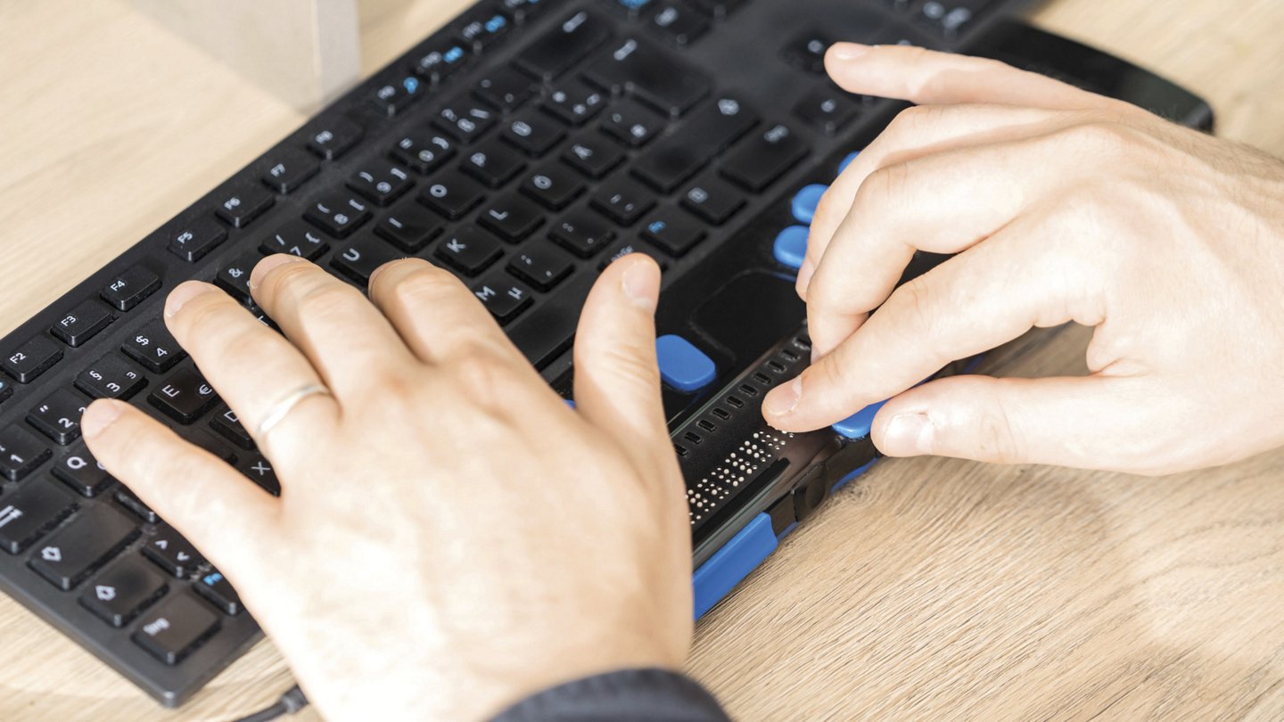 Hände einer Person arbeiten an einer Tastatur mit taktilen und technischen Hilfsmitteln für barrierefreie Bedienung.