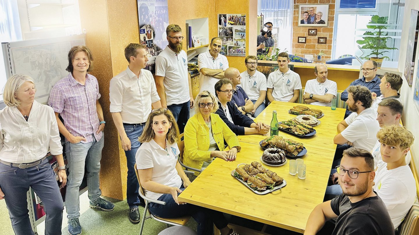 Eine Gruppe von etwa 15 Personen sitzt und steht um einen Tisch in einem Büro oder Besprechungsraum. Der Tisch ist mit Snacks und Getränken gedeckt. Die Gruppe besteht aus Männern und Frauen in unterschiedlichem Alter, einige tragen Firmenkleidung. Die Atmosphäre wirkt entspannt und gesellig.