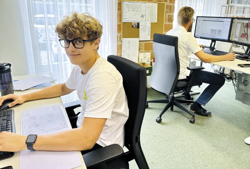 Zwei Männer sitzen an Computern in einem Büro. Einer der beiden, ein junger Mann mit lockigem Haar und Brille, schaut lächelnd in die Kamera. Der zweite Mann arbeitet konzentriert an seinem Bildschirm. Auf den Monitoren sind technische Zeichnungen zu sehen.