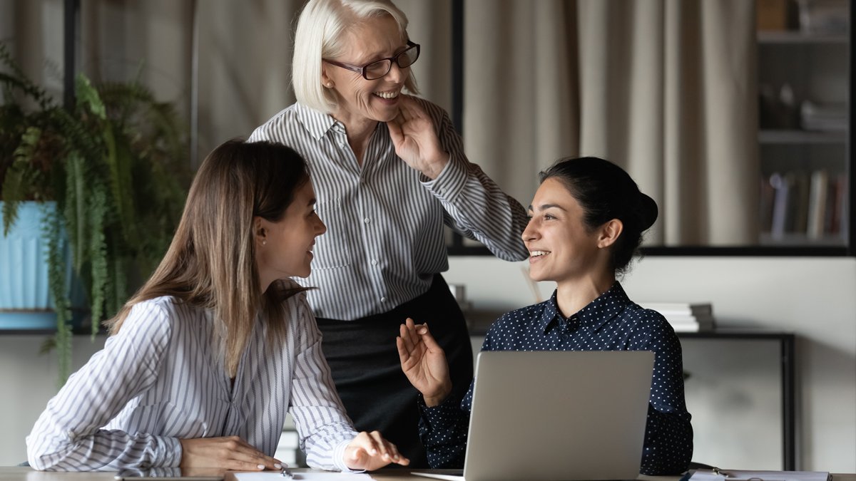 Drei Frauen am Arbeitsplatz. Sie besprechen etwas vor einem offenen Laptop. Im Vordergrund sitzen zwei jüngere während eine ältere Frau hinter ihnen steht und lächelt.