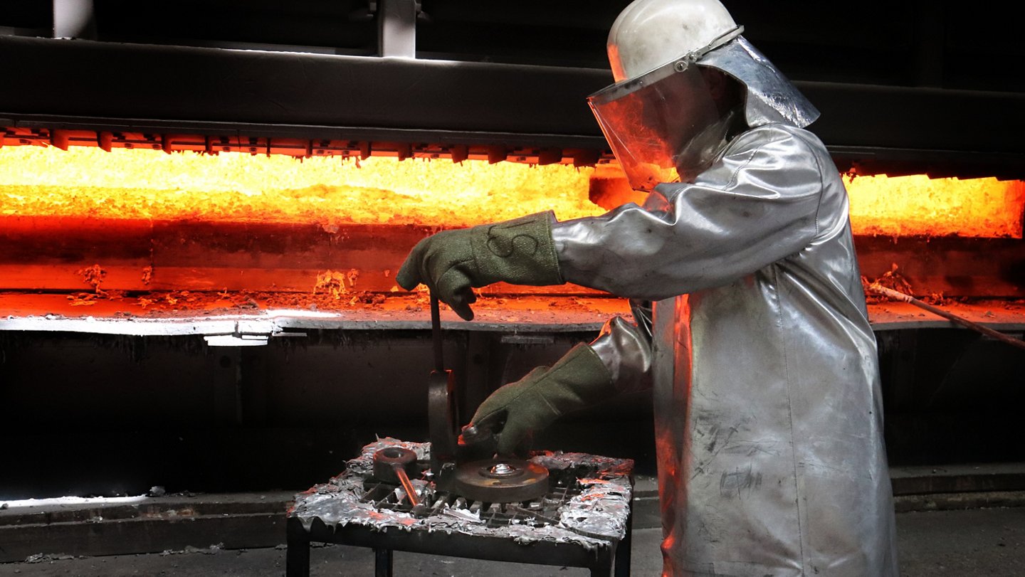 Nahaufnahme Arbeiter im Schutzanzug untersucht vor dem Hochofen eine Probe flüssigen Aluminiums.