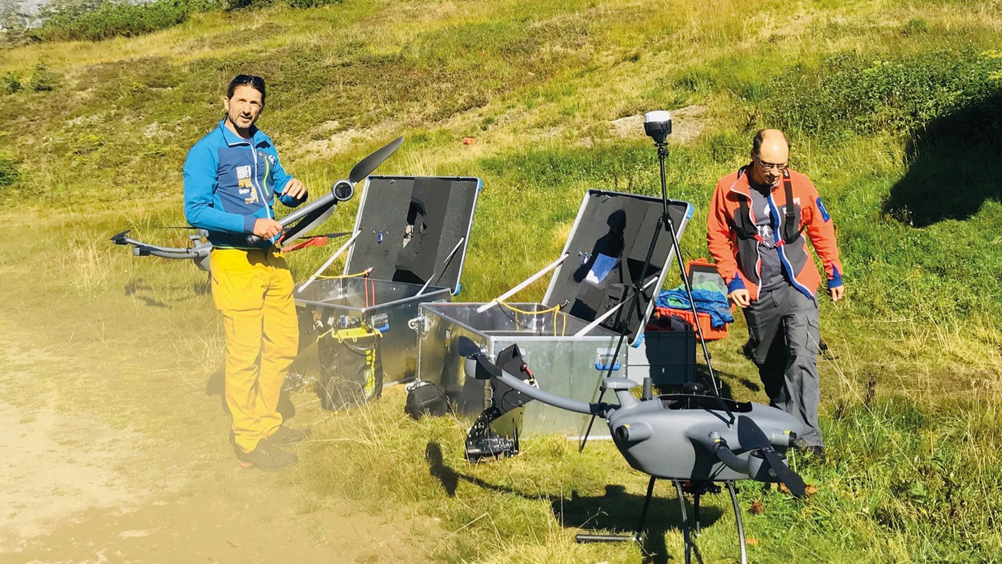 zwei Männer mit zahlreichen Geräten, großen Metallkisten und Drohnen in einer Berglandschaft
