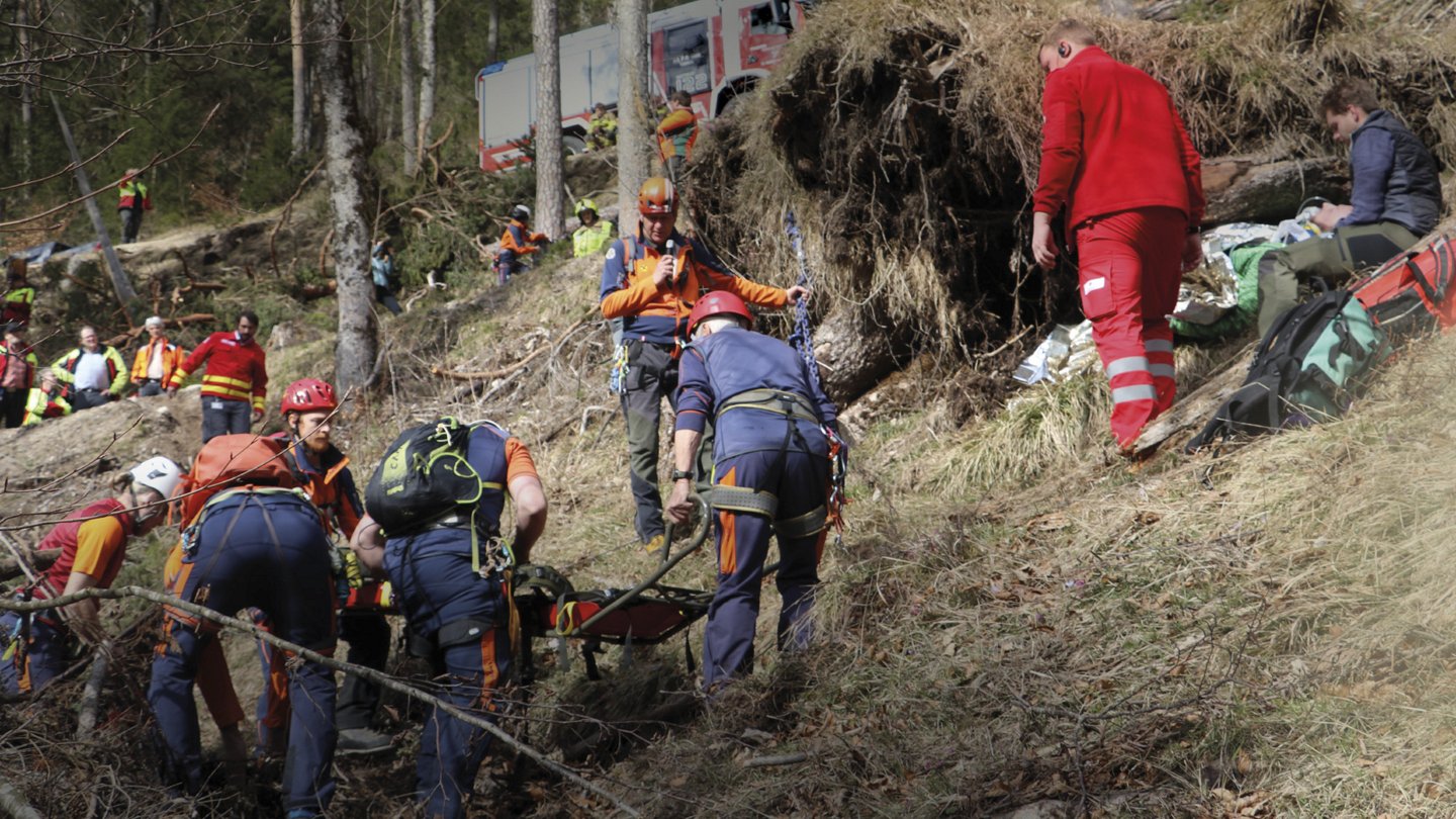 Ein Rettungsteam arbeitet an einem Berghang, umgeben von umgestürzten Bäumen und Vegetation. Im Hintergrund sind Rettungsfahrzeuge und weitere Einsatzkräfte zu sehen. Die Rettungskräfte bereiten die Bergung eines verletzten oder eingeschlossenen Individuums vor.