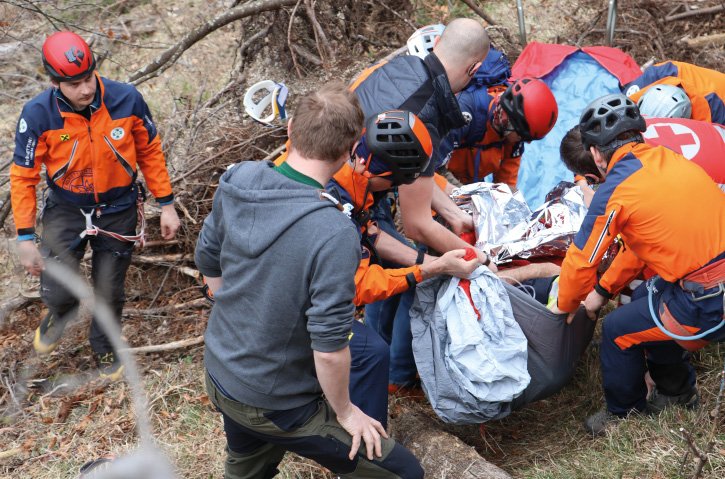 Mehrere Rettungskräfte in orangefarbener Schutzkleidung und Helmen bereiten sich darauf vor, eine verletzte Person in einer Trage, die in eine Rettungsdecke eingewickelt ist, den Hang hinunter zu transportieren.