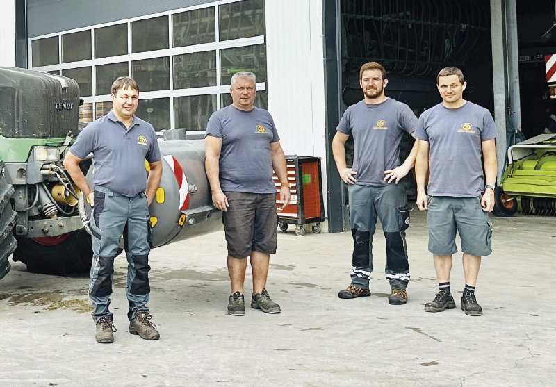  Eine Gruppe von fünf Männern steht vor großen landwirtschaftlichen Maschinen, darunter ein grüner Traktor und eine gelbe Ballenpresse, vor einem Gebäude mit großen Toren. Die Männer sind in Arbeitskleidung gekleidet und lächeln in die Kamera.