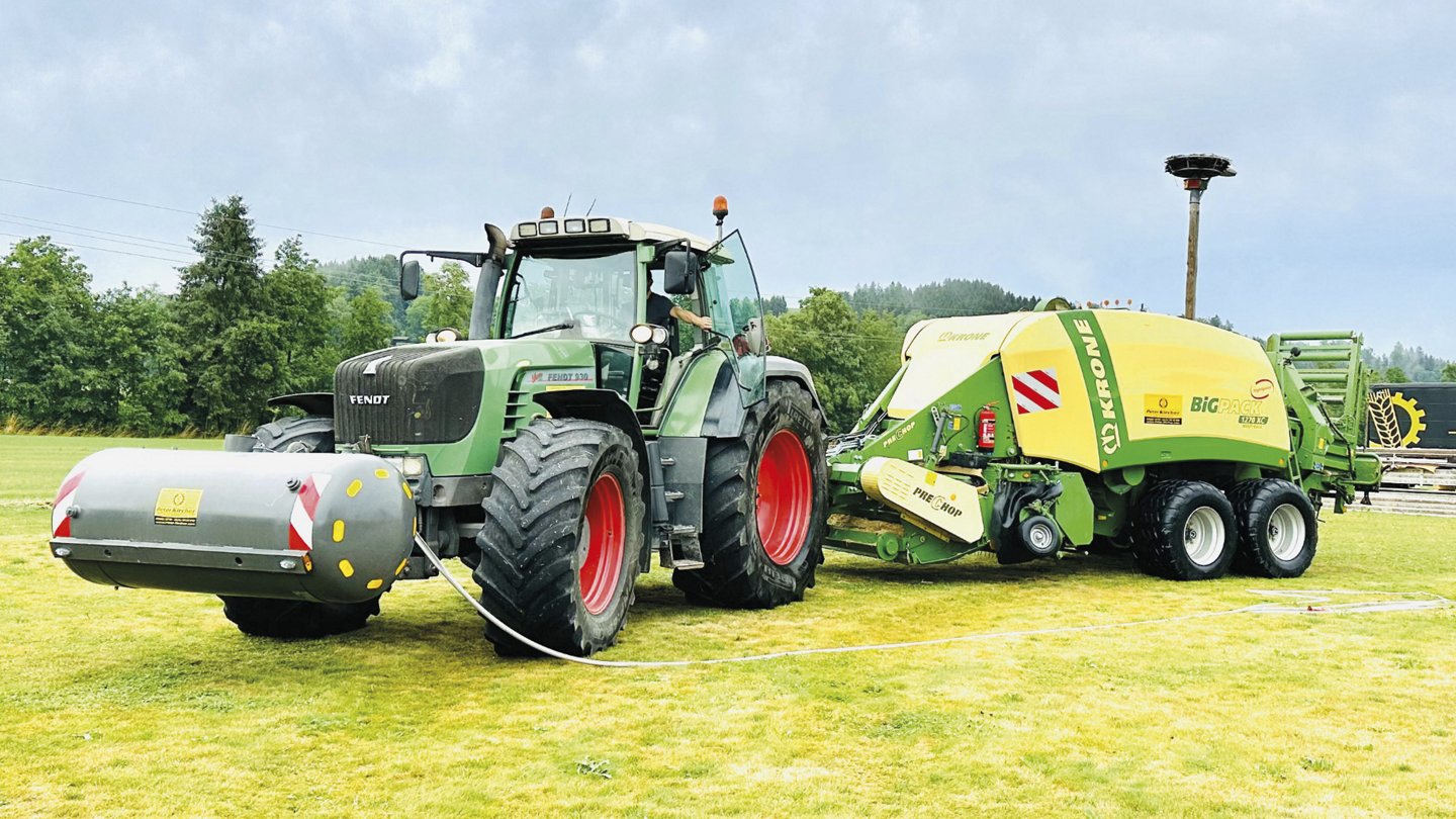 Ein grüner Traktor ist auf einer Wiese mit einer gelben Ballenpresse verbunden. Im Hintergrund ist ein Schild mit der Aufschrift "Peter Kircher" und dem Slogan des Unternehmens zu sehen. Der Himmel ist bewölkt.