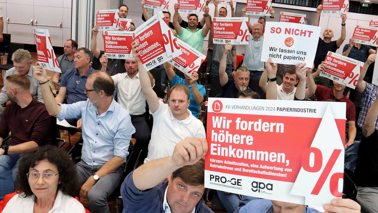 Konferenzteilnehmer:innen mit Schildern "Wir fordern höhere Einkommen" und "Wir lassen uns nicht papierln"