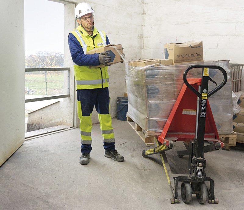 Ein Arbeiter in Schutzkleidung und Helm hebt einen Karton auf einen Hubwagen, darauf befinden sich viele weitere Kartons.