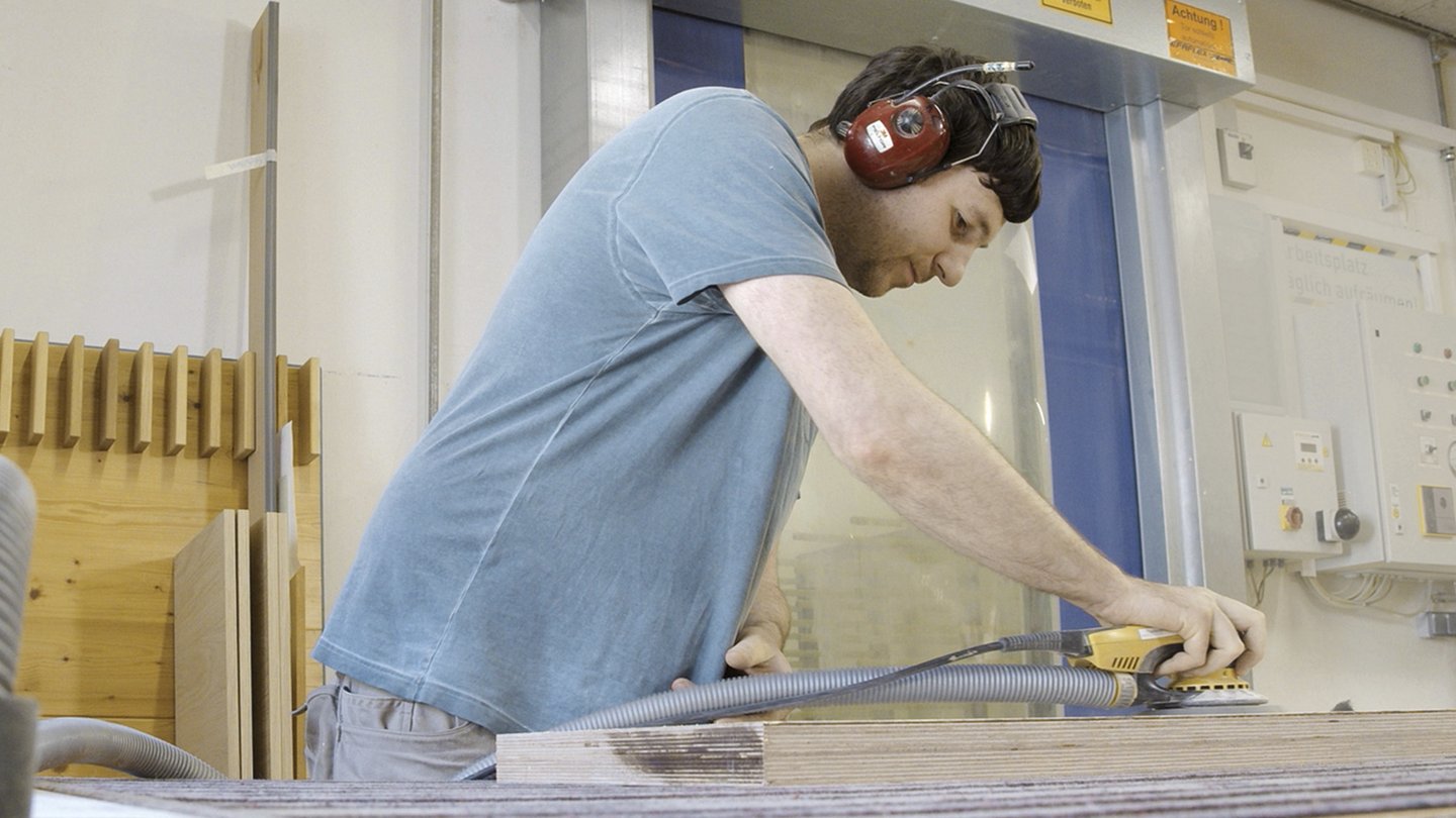 ein Mann mit Hörschutz arbeitet an einem Stück Holz mit einer Schleifmaschine