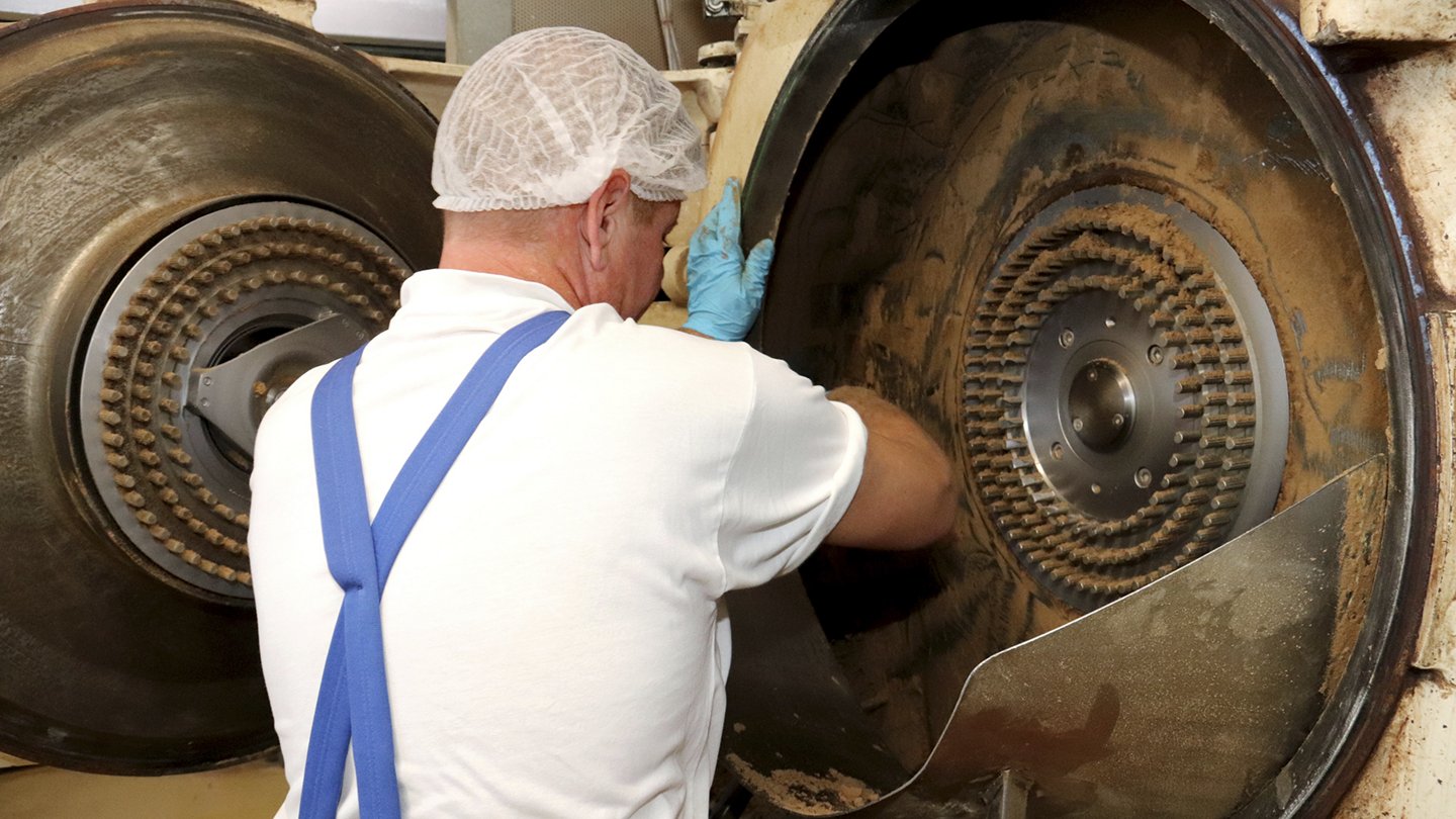 Ein Mann schabt Gewürzreste aus einer großen, aufgeklappten Stiftmühle.