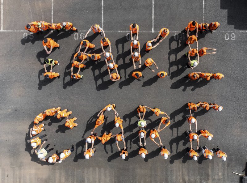 Das Bild zeigt eine Gruppe von Menschen in orangener Arbeitskleidung und Helmen, die auf einem asphaltierten Platz in Form der Worte „TAKE CARE“ posiert haben. Die Anordnung wurde aus der Vogelperspektive aufgenommen.