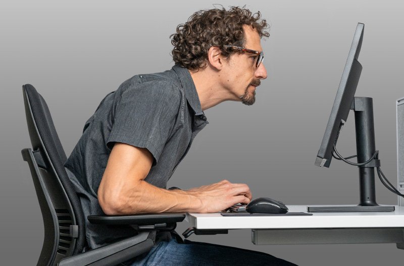 Schlechte Körperhaltung durch Lesebrille: Ein Mann beugt sich nach vorne, um Inhalte auf dem Monitor durch eine ungeeignete Lesebrille scharf zu sehen, was eine unergonomische Haltung verursacht.