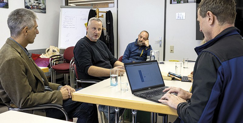 mehrere Männer sitzen an einem Tisch und besprechen etwas, ein Mann tippt in sein Notebook