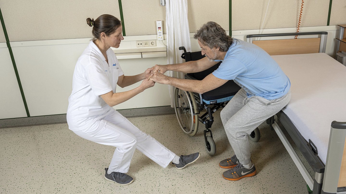 Pflegekraft hilft einer Patientin im Krankenhausbett, sich aufzusetzen, um den Transfer in einen Rollstuhl vorzubereiten. Die Patientin greift nach den Händen der Pflegekraft zur Unterstützung.