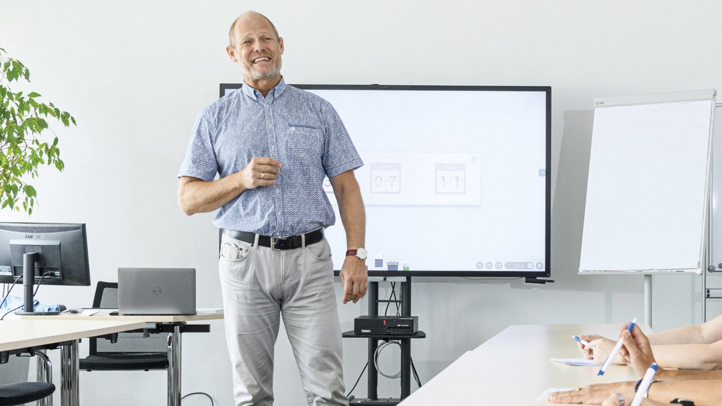 Das Bild zeigt eine Schulungssituation in einem modernen Konferenzraum. Der Dozent, **Christian Schenk**, Schulungsleiter Prävention in der **AUVA-Hauptstelle**, steht vor einer digitalen Präsentation auf einem großen Bildschirm. Die Teilnehmer:innen, deren Hände im Vordergrund sichtbar sind, machen sich Notizen.   Die Atmosphäre wirkt professionell und freundlich. Die **Schulungsplattform AUVkurs.at** wird oben im Bild erwähnt, was darauf hindeutet, dass es sich um ein offizielles Weiterbildungsangebot handelt.   Das Setting beinhaltet moderne Technik wie einen Bildschirm mit interaktiver Präsentation, Laptops und einen Flipchart, was die digitale und praxisnahe Ausrichtung der Schulung unterstreicht.