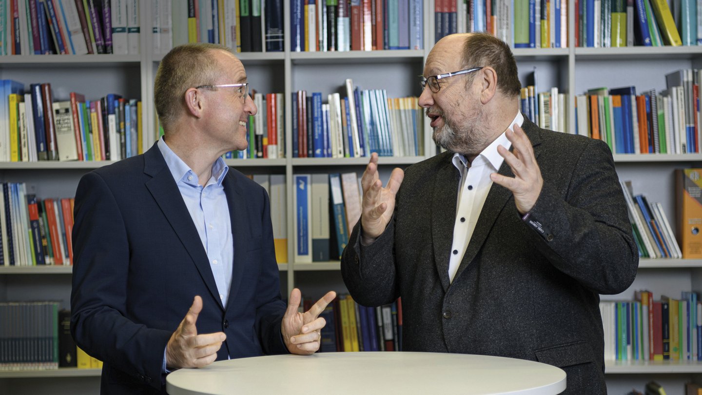 zwei Männer besprechen sich, dahinter ein Bücherregal