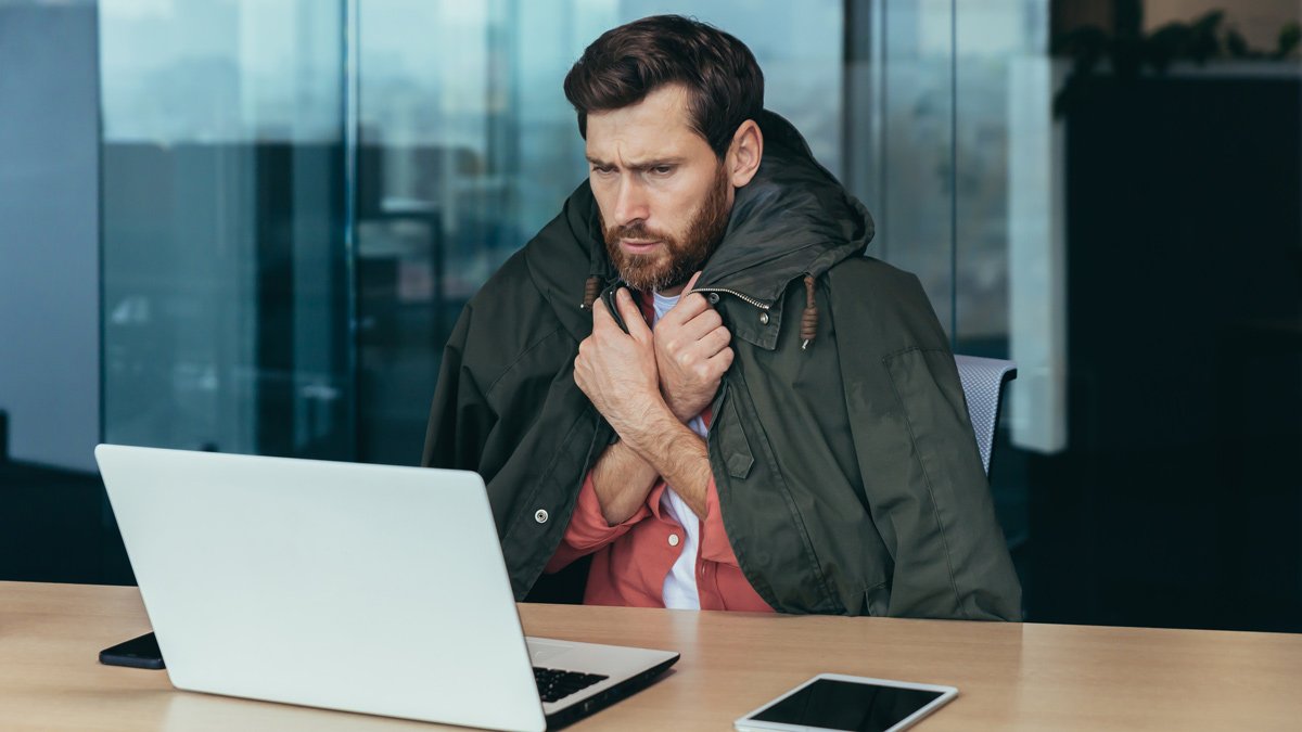 Mann sitzt, mit übergeworfener Jacke, frierend im Büro.