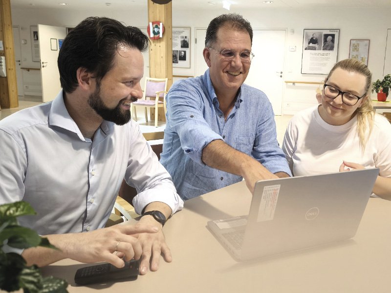 Zwei Männer und eine Frau sitzen an einem Tisch, schauen auf einen Laptop und diskutieren lächelnd in einer freundlich gestalteten Umgebung.