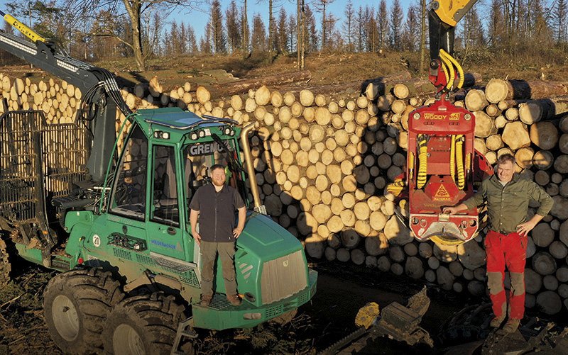 zwei Männer stehen vor einem Stapel Holz und großen Forstmaschinen