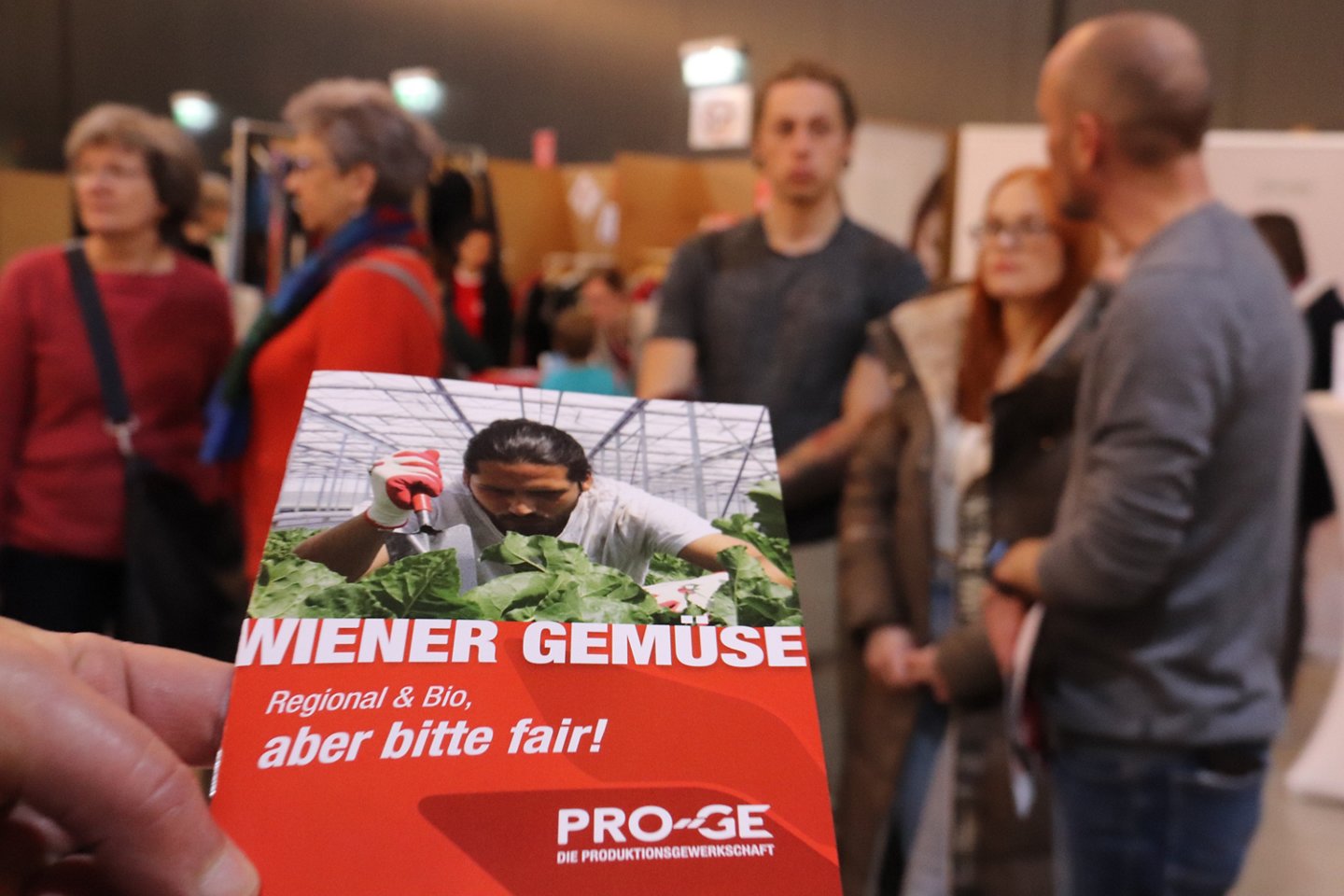 Folder im Vordergrund, im Hintergrund unscharf Menschen beim PRO-GE Stand